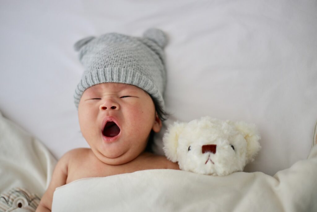 baby in gray knit hat yawning while tucked into bed with a cute teddy bear