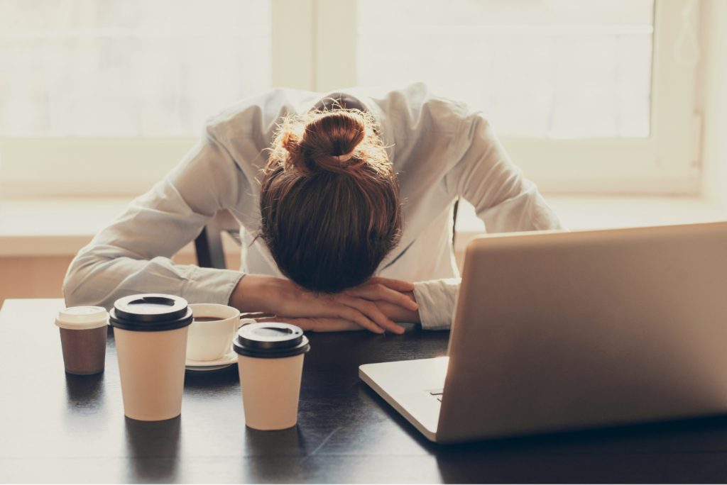 a woman sits at a desk in front of a laptop surrounded by 4 coffee cups. She rests her head against the desk tiredly. 