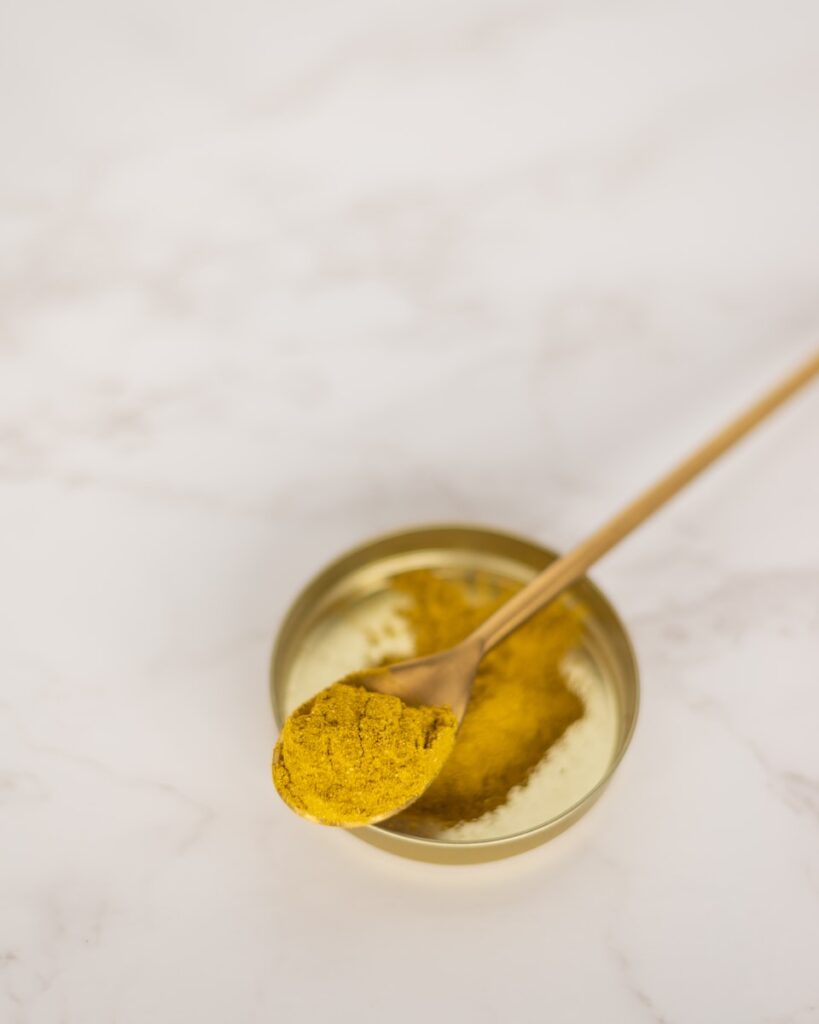 stainless steel spoon on clear glass bowl with yellow liquid & the spoon is full of turmeric powder