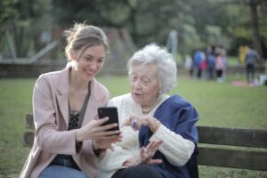 adult granddaughter shows her elderly grandmother something on her phone sitting on a park bench.