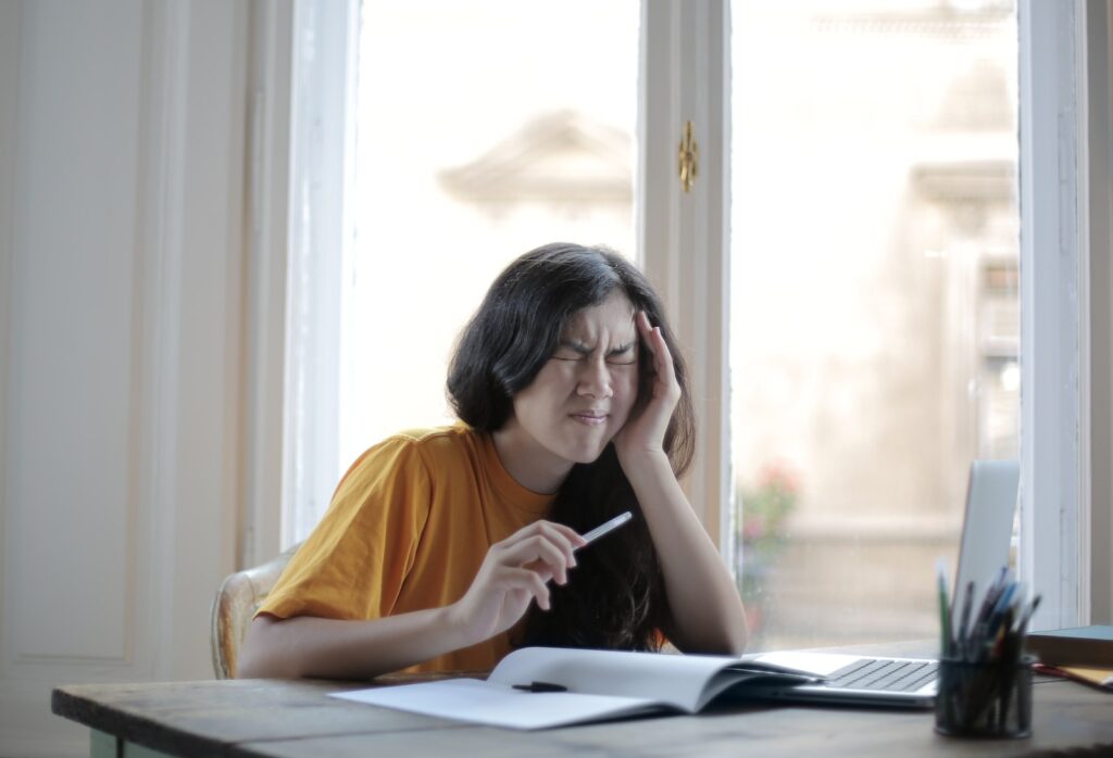  female student in casual wear sitting at table with laptop and notebook and grimacing from migraine while doing homework during distance learning