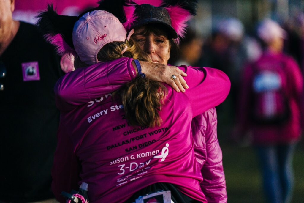 a woman in a pink shirt hugging another woman emotionally at a breast cancer fundraising event