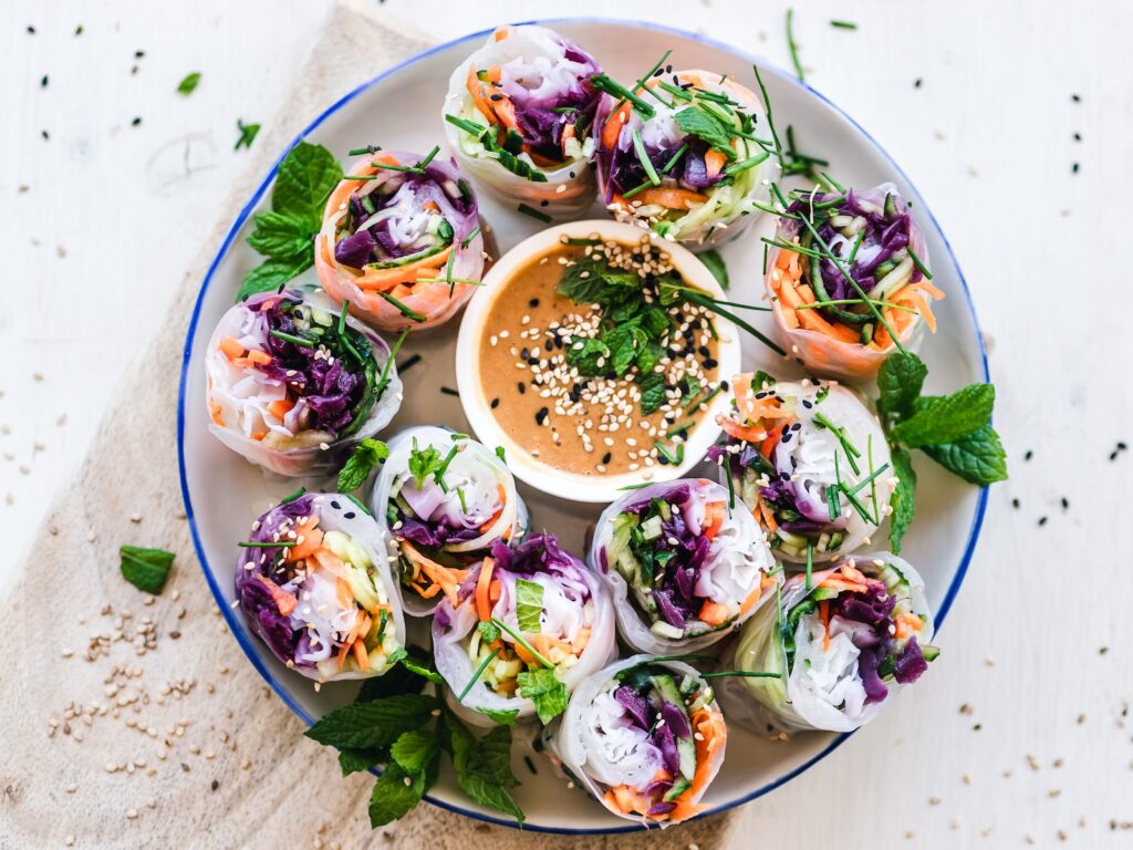 vegetable summer rolls in rice paper with colorful vegetables inside, surrounding a bowl of peanut sauce.