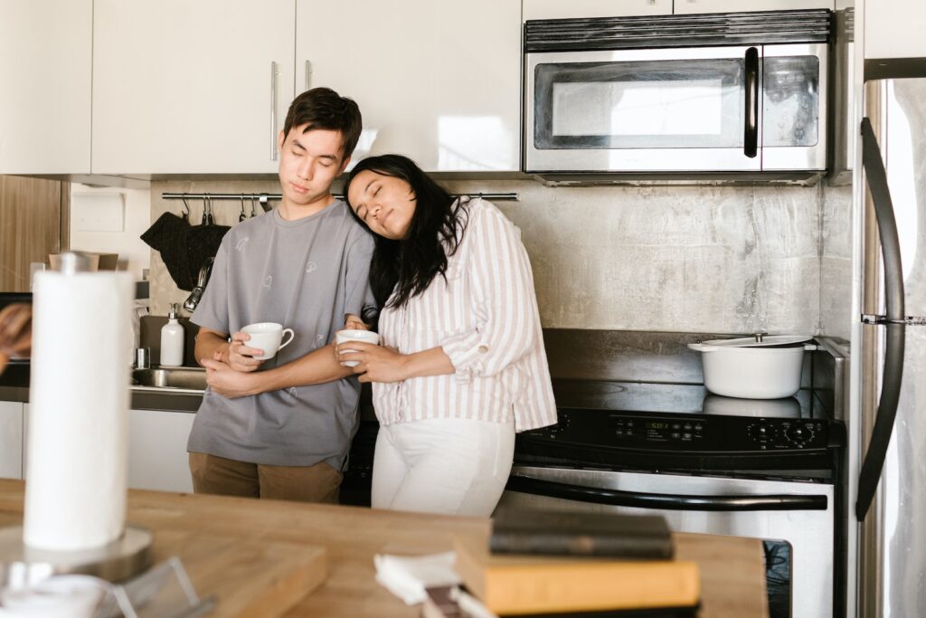 a couple standing in their kitchen looking tired, holding cups of coffee