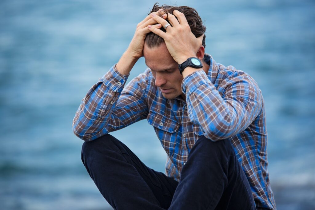 Man in Blue and Brown Plaid Dress Shirt sitting outside looking distressed, holding his head in worry