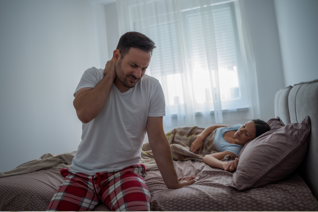 a man sitting on the edge of his bed grasping his neck in pain while his wife sleeps