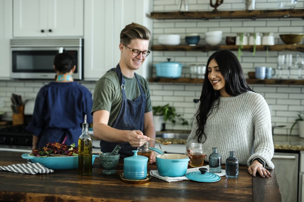 blonde man and brunette woman cooking together in white kitchen with wood accents and blue cookware 