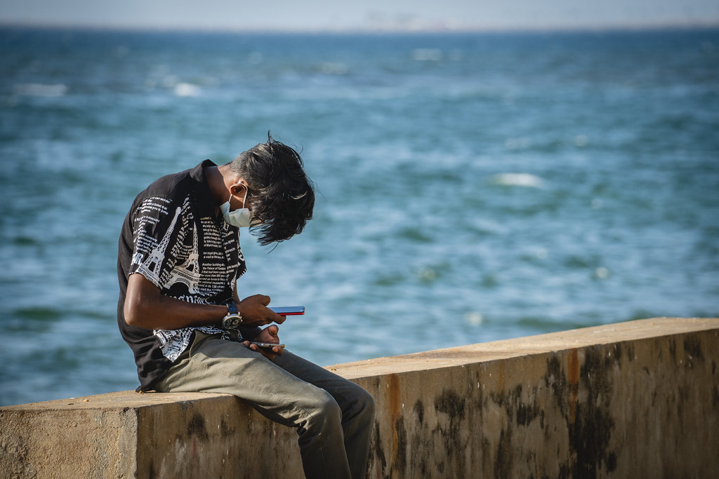 a person sits on a wall overlooking the ocean hunched over their cell phone 