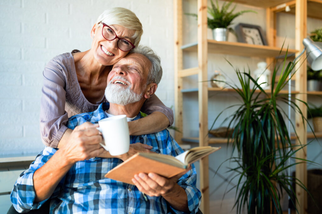 an older couple embracing. The husband is sitting down reading a book and drinking coffee, while the wife is hugging him from behind. 