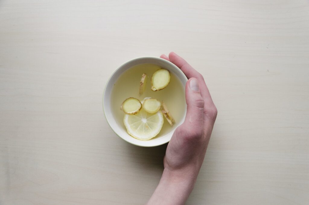 person holding white teacup with tea, sliced lemon and ginger inside