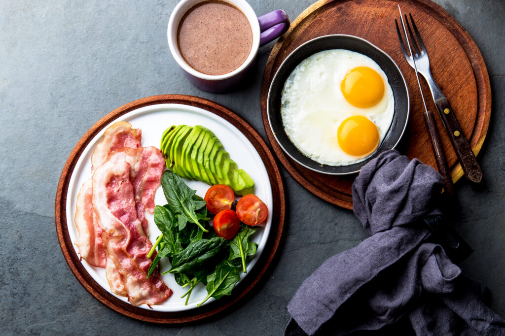 A keto diet breakfast: sunny side up eggs with bacon, avocado, tomatoes and some greens. There's also a black coffee. 