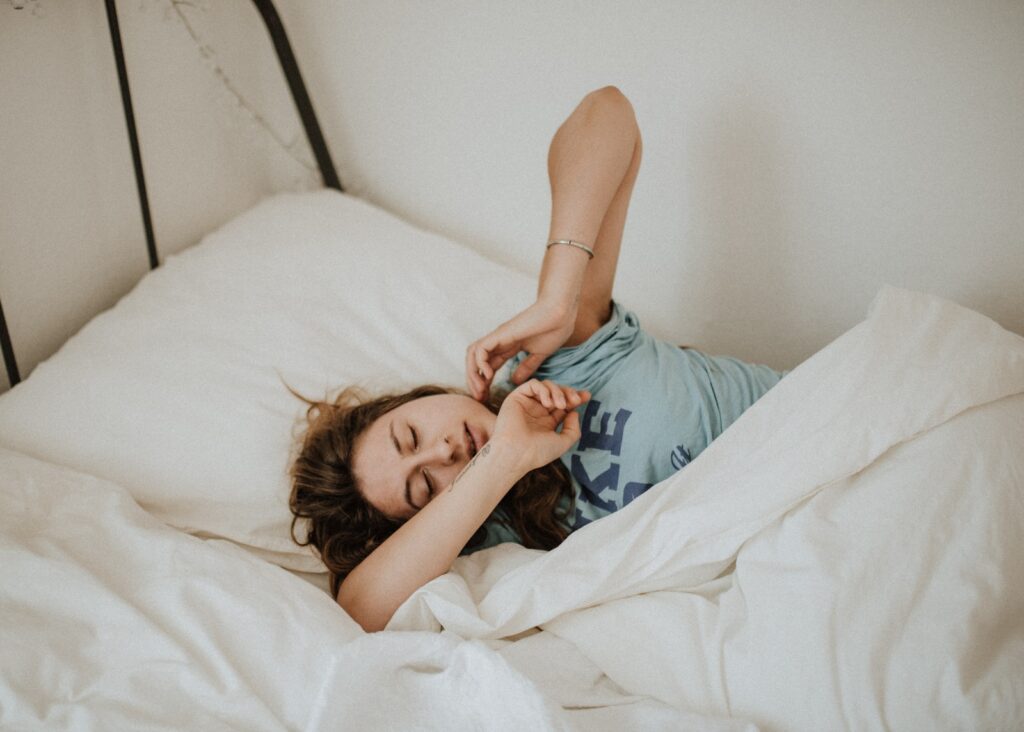 woman covered in white blanket sleeping on white bed comforter stretching her arms out