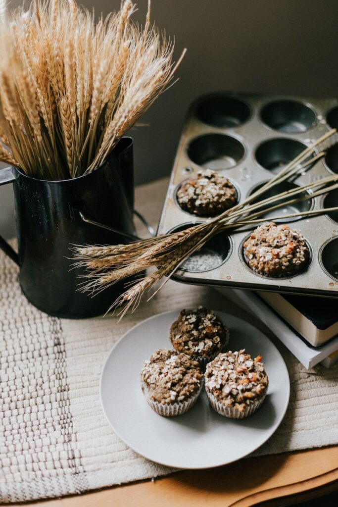 whole grain muffins in the muffin tin beside a black pitcher full of wheat. 