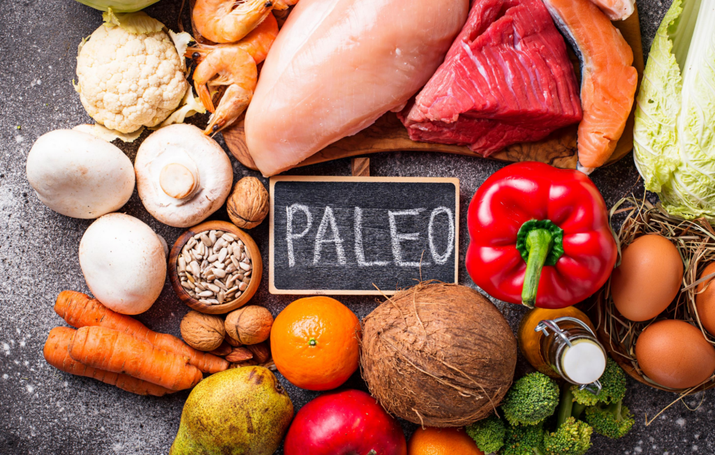 a variety of paleo ingredients surrounding a small chalkboard that says "Paleo". There are different types of meat, fish, eggs, vegetables, and nuts. 