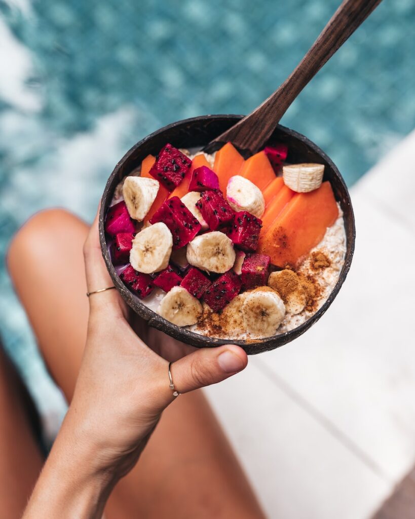 a woman holding a bowl of fruit and oatmeal while sitting on the side of a pool. 