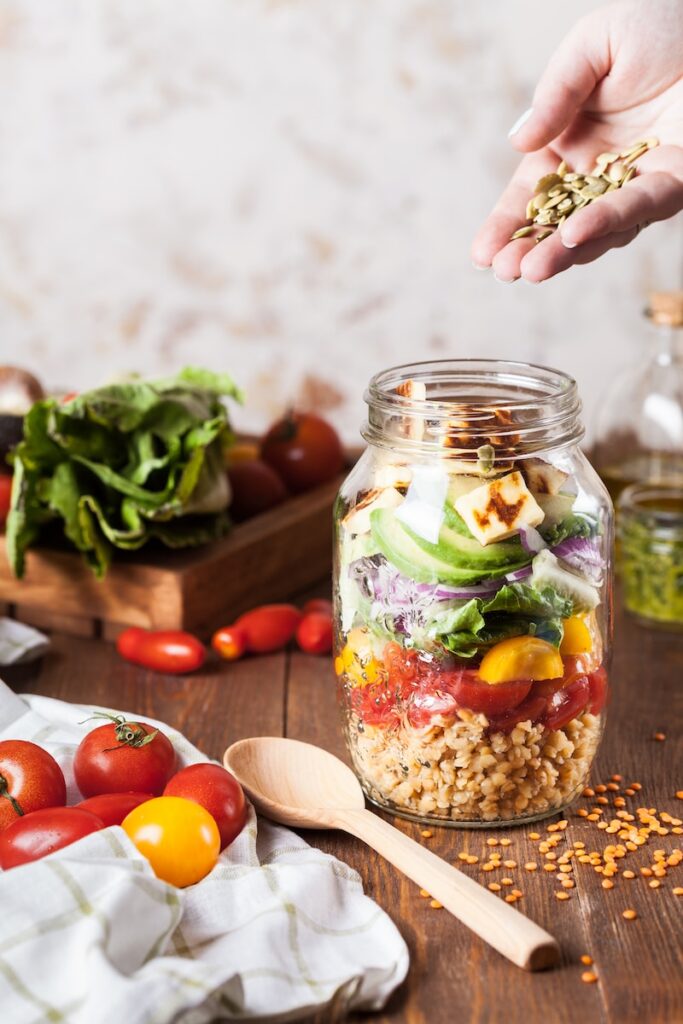 someone adding garnishes to a jar salad that has barley, veggies, and tofu in it. 