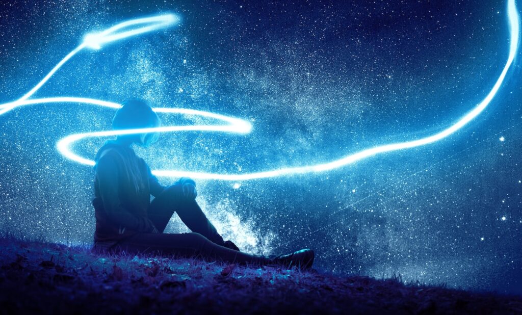 dream-like photo of a person sitting on blue grass with a streak of blue light circling them and the starry night sky behind them. 