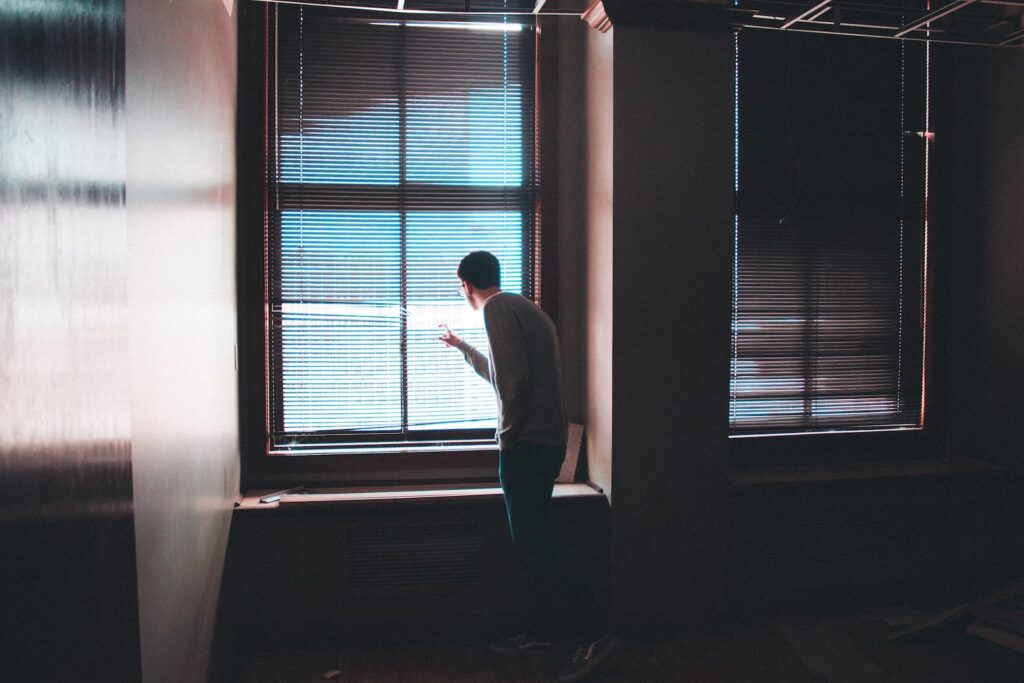 man in the dark peeking out the blinds covering a window 